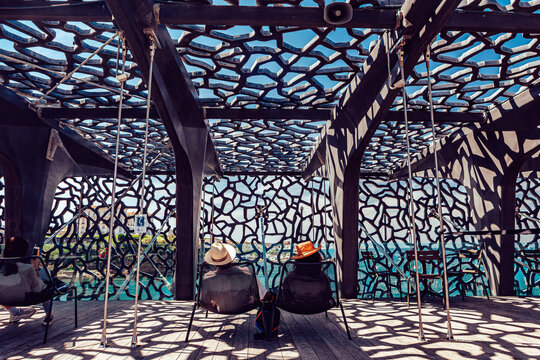 View of a captivating dance of light and shadow as two figures sit beneath a modern, organic-patterned structure overlooking the shimmering sea, Marseille, Provence-Alpes-Cote d'Azur, France.