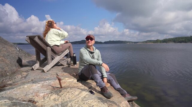 Relaxed mature couple enjoying sun on rocky archipelago coast. Authentic slow living, mental wellness, and sustainable travel concept in Nordic nature landscape.
