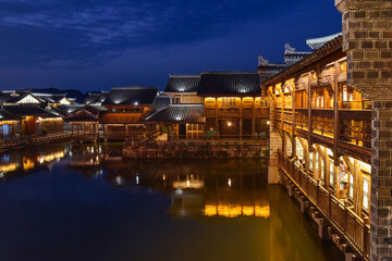 Fototapeta premium Night view of traditional stilt houses with glowing lanterns reflecting on the river in Moon Fields (Yueliangtian) scenic spot in Lizhuang Ancient Town, Yibin, Sichuan, China