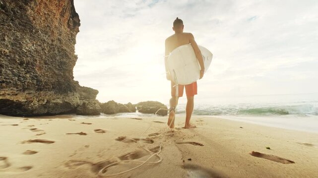 Surfer walks along a sandy beach toward the ocean waves at sunset, carrying a surfboard under his arm. The warm golden light illuminates the scene, with a dramatic rocky cliff in the background and