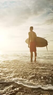 Young surfer stands on the beach at sunset, holding his surfboard near a majestic cliff. Warm backlight from the sun illuminates the scene, with sparkling ocean waves in the background, creating a