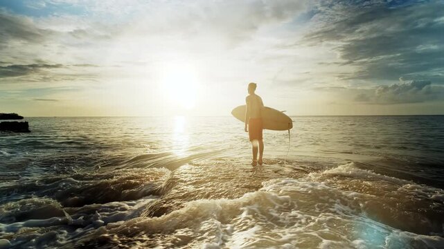 Young surfer stands in the ocean at sunset, holding his surfboard and savoring a tranquil moment before catching the waves. Warm golden hour light reflects off the water, highlighting the serene