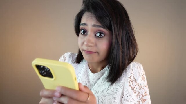 Close up of confused cute young Asian girl woman using browsing smart phone receiving unknown sms messages, looking at camera nodding head negative reaction, isolated over grey studio background.