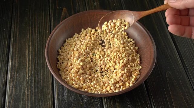 A hand pours yellow split peas from a wooden spoon into a clay bowl, demonstrating continuous movement in a rustic kitchen environment. Preparing to cook a pea dish