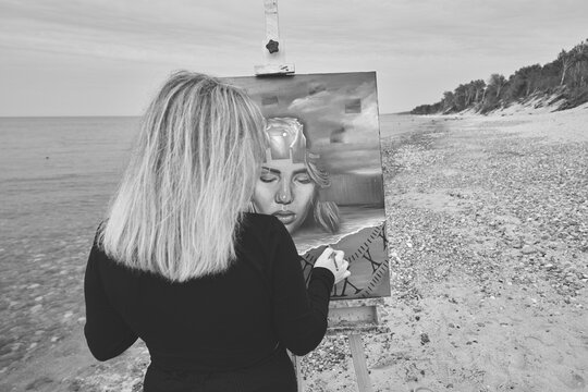 Young woman painting self-portrait on easel at peaceful beach setting
