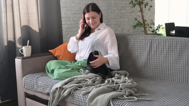 Woman sitting on sofa drinking coffee, using smartphone, and petting griffon dog. Concept of relaxation, anti stress lifestyle, cozy home atmosphere, and living with pets.