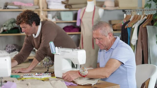 Fashion designer draws a sketch and a size grid on paper, next to a male tailor who works on a sewing machine. Men create a collection of clothes in a sewing workshop. High quality 4k footage