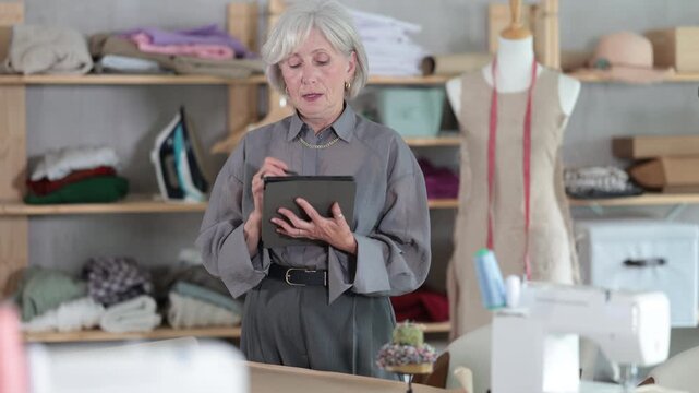 Mature dressmaker draws a sketch of clothes and patterns on an electronic tablet. Woman creates a fashion design in a workshop.