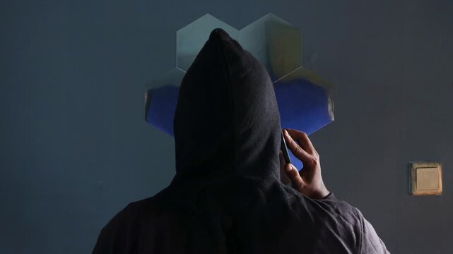 Anonymous spy person or hacker with black hoodie talking on the phone indoors. Standing near a mirror in the room. Mysterious man making phone call conversation with his back to camera.
