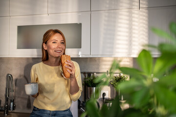 Woman enjoying breakfast sandwich and coffee in kitchen