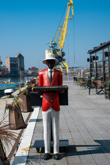 a statue depicting a waiter in a red jacket, white hat, and white trousers holding a tray, standing on a paved waterfront promenade with a large yellow harbor crane and outdoor cafe seating 