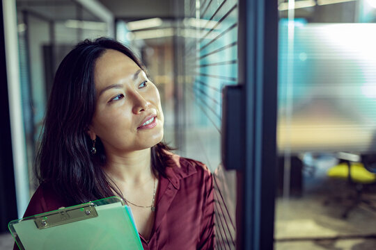 Thoughtful businesswoman with clipboard by glass wall in modern office