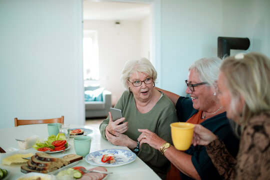 Senior women friends sharing coffee and smartphone at home kitchen