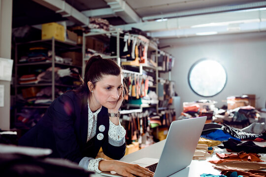 Female entrepreneur working on laptop in fashion studio stockroom