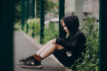 A thoughtful sad young boy sits alone by a green fence wearing a black hoodie and worried about his problems