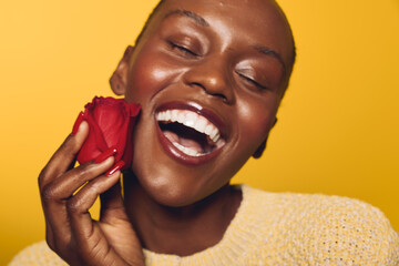 Beautiful dark skinned African woman with closed eyes smiling widely holds red rose near face on...