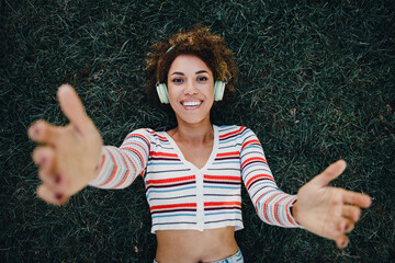 Playful woman with headphones lying on grass reaching out smiling during a sunny outdoor moment