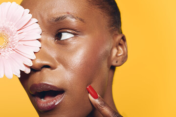 Close-up of a dark skinned African woman with natural skin texture holding a pink flower near her face on yellow background. Concept of beauty, skincare, natural cosmetics, and wellness.