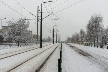 Tram tracks and a pedestrian path stretch through a snowy urban corridor lined with leafless trees and utility poles. Minimal winter transport scene conveys cold weather, commuting infrastructure, and