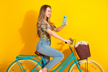 Young stylish woman rides a teal bike while taking a selfie against a bright yellow background