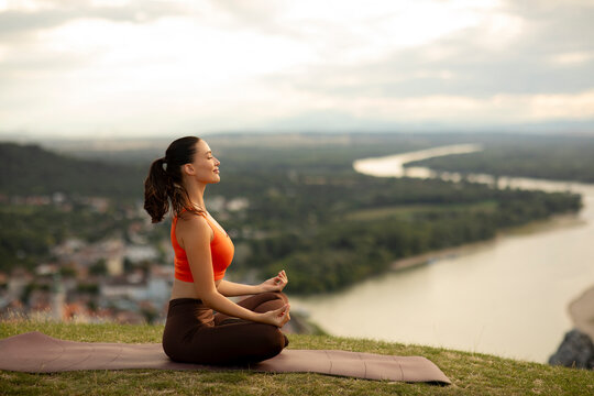 Side view of young woman sitting cross-legged on mat, focusing on her breath and practicing yoga outdoors, with river flowing on background