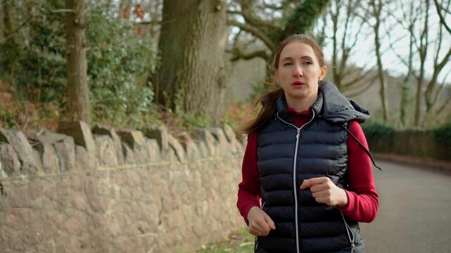 Jogging woman moving along stone wall pathway in wooded park during daytime workout. Female runner training outdoors building stamina and improving cardiovascular health. Active fitness routine