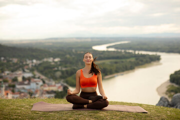 Fototapeta premium Lady sitting on mat, meditating and practicing yoga beside river surrounded by hills and city in the background. Mindful mountain escape