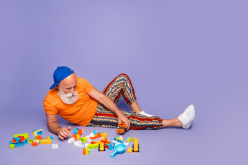 Fototapeta premium Older man in orange tshirt and striped pants plays with colorful blocks on a purple violet background