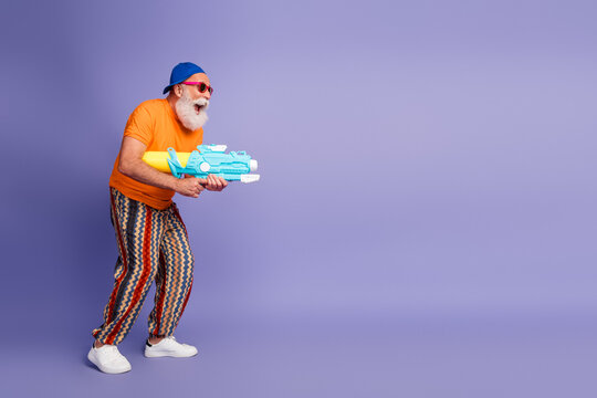 Old man with bright orange shirt and striped pants aims a blue toy water gun against a purple background