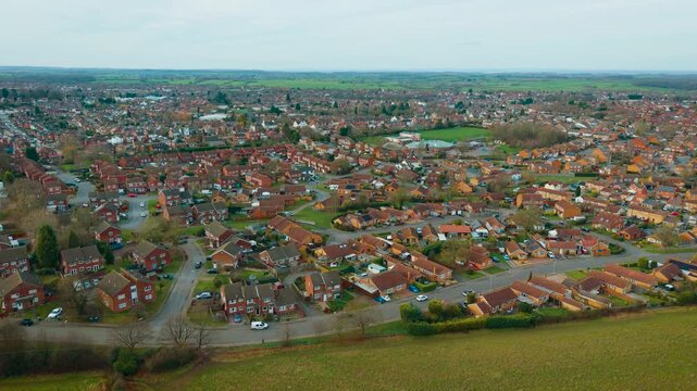 Suburban housing estate aerial panorama with red brick homes and winding residential roads. Compact neighborhood layout bordering green agricultural fields and rolling countryside. Broad town