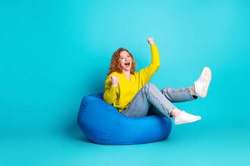 Cheerful young woman with curly red hair wearing a yellow sweater sits on a blue bean bag...