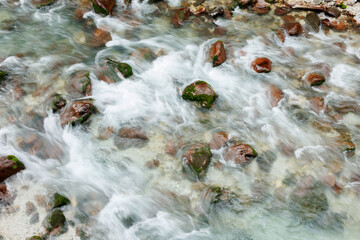 A dynamic top-down perspective of a clear mountain stream in the Julian Alps, captured with a slow shutter speed to create a silky white motion blur as water cascades over reddish-brown, moss-covered  © Timur Abasov