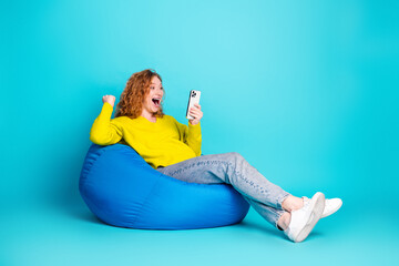 Fototapeta premium Young woman in a bright yellow sweater relaxing on a blue beanbag smiles at her phone in a vibrant teal studio background