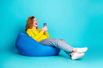 Young woman with red hair sits on a blue bean bag smiling at her phone against a bright teal...