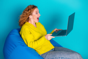 Surprised young woman wearing a yellow sweater using laptop while sitting on a blue bean bag