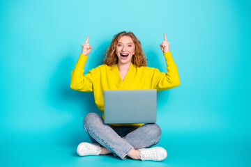 Young woman with red hair wearing a bright yellow sweater sits cross legged with a laptop...