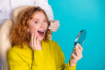 Young redhead woman smiles during dental checkup in bright yellow sweater with mirror on teal...