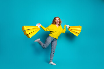 Energetic young woman with yellow sweater jumps with shopping bags against teal blue background