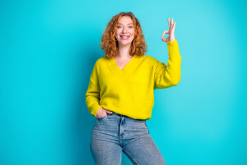Young woman with curly red hair in a bright yellow sweater smiles and makes an ok gesture against a...