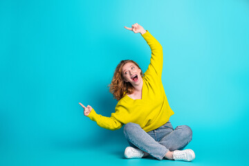 Young woman in yellow sweater sits on blue background smiling and pointing excitedly