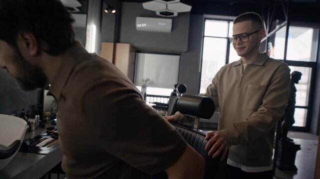 Handheld shot of two men shaking hands in barbershop. Man sitting in chair and explaining his preference while barber listening to him carefully