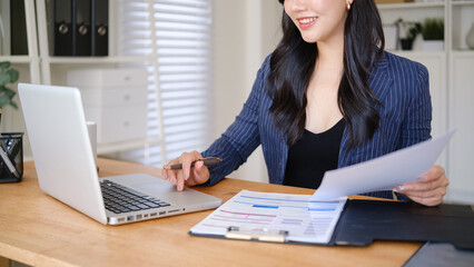 Close up of businesswoman reviewing financial documents while using a laptop at a modern office...