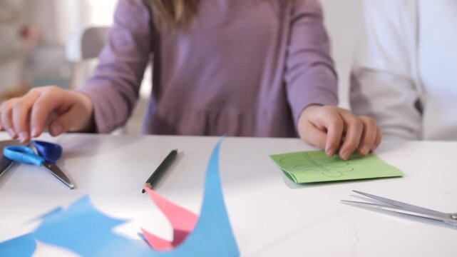 Child's hands cutting and drawing colorful paper shapes with scissors and pencil at a table, focused on a simple arts and crafts project to develop fine motor skills and creativity