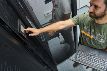 It technician adjusting server control panel in a data center while holding a laptop for diagnostics