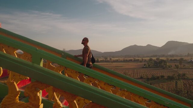 Visitors walk along a colorful walkway toward Wat Tham Suea, the famous Tiger Cave Temple in Kanchanaburi, Thailand, featuring ornate golden Buddhist architecture on a hilltop.