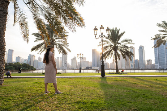 Young woman enjoying sunset view of Sharjah Corniche, female tourist looking at city skyline with skyscrapers and palm trees over Khalid Lagoon in United Arab Emirates at twilight.