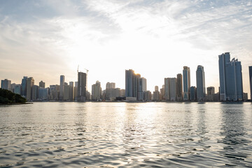 Obraz premium 12.12.2025 Sharjah, UAE. Panoramic view of Sharjah Corniche at sunset, featuring Khalid Lagoon water, illuminated skyscrapers, modern city skyline, and silhouette of date palm trees in UAE