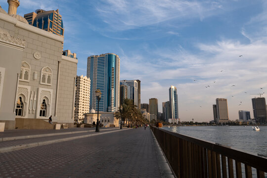 12.12.2025 Sharjah, UAE. Panoramic view of Sharjah Corniche at sunset, featuring Khalid Lagoon water, illuminated skyscrapers, modern city skyline, and silhouette of date palm trees in UAE