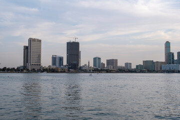 Fototapeta premium 12.12.2025 Sharjah, UAE. Panoramic view of Sharjah Corniche at sunset, featuring Khalid Lagoon water, illuminated skyscrapers, modern city skyline, and silhouette of date palm trees in UAE