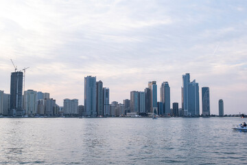 Fototapeta premium 12.12.2025 Sharjah, UAE. Panoramic view of Sharjah Corniche at sunset, featuring Khalid Lagoon water, illuminated skyscrapers, modern city skyline, and silhouette of date palm trees in UAE